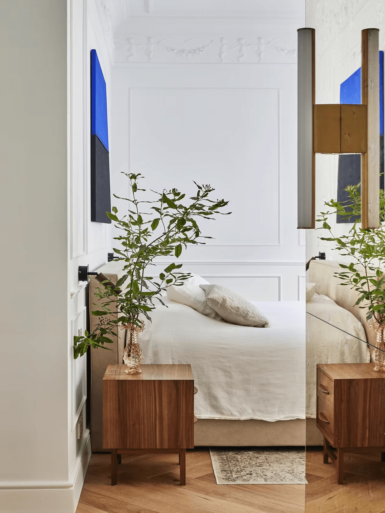 Bedroom with white linens, mirrored wall, walnut nightstand, and brass lighting